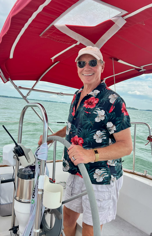 A person wearing sunglasses, a floral shirt, and a cap is standing at the helm of a boat with a red canopy, surrounded by water.