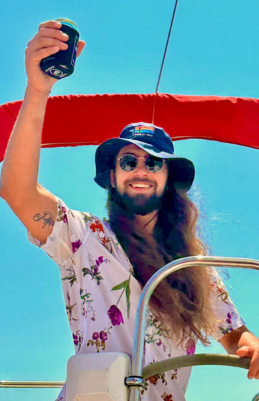 Man with long hair and beard wearing a floral shirt and hat, holding a drink can, standing on a boat with a red canopy against a clear blue sky.