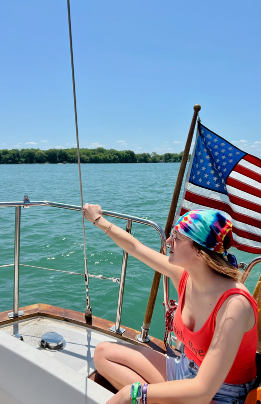 Person wearing a colorful headscarf and red tank top sitting on a boat, with an American flag in the background and water and trees visible.