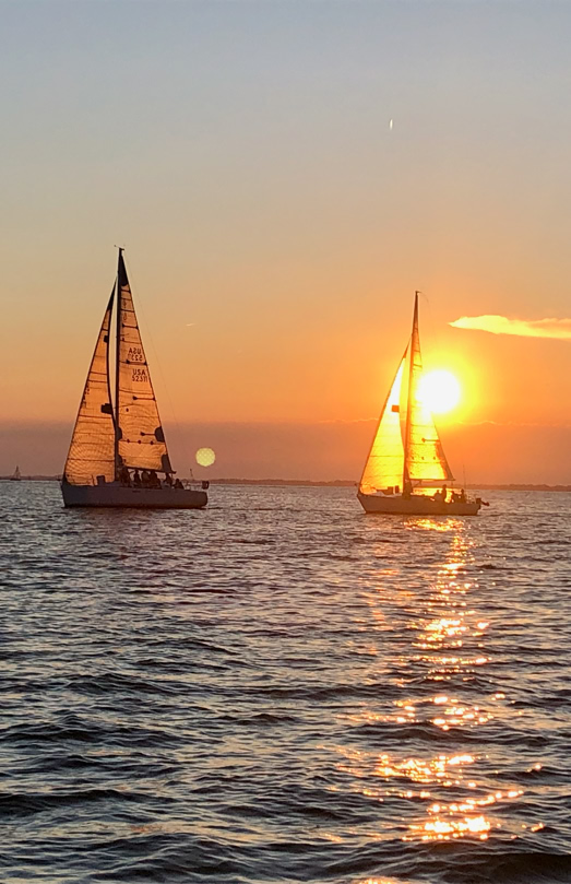 Two sailboats on the water at sunset with an orange sky and the sun near the horizon.