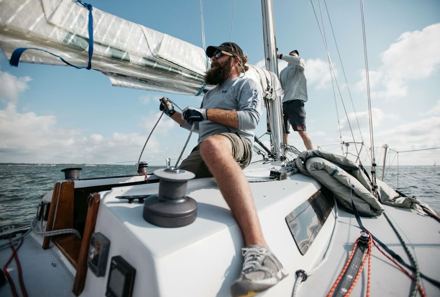 Two people on a sailboat, one sitting and handling ropes, the other standing near the mast, with the sea and sky in the background.