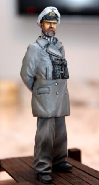 A small figurine of a person in a gray military uniform with a cap, standing with hands behind their back, and binoculars hanging from the neck.