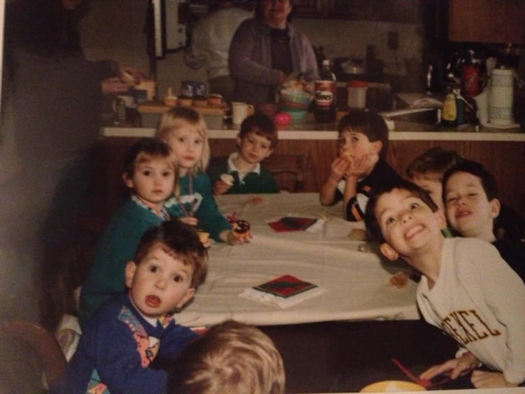 A group of children sitting around a table with a white tablecloth, some looking at the camera, in a kitchen setting with an adult in the background preparing food.