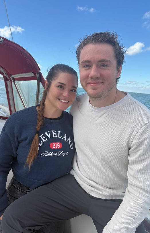 A man and woman sitting together on a boat with a blue sky and ocean in the background. The woman is wearing a navy sweatshirt, and the man is wearing a white long-sleeve shirt.